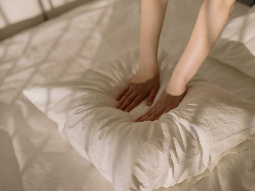 A warm, sunlit bedroom scene with hands fluffing a soft pillow, creating a cozy atmosphere.